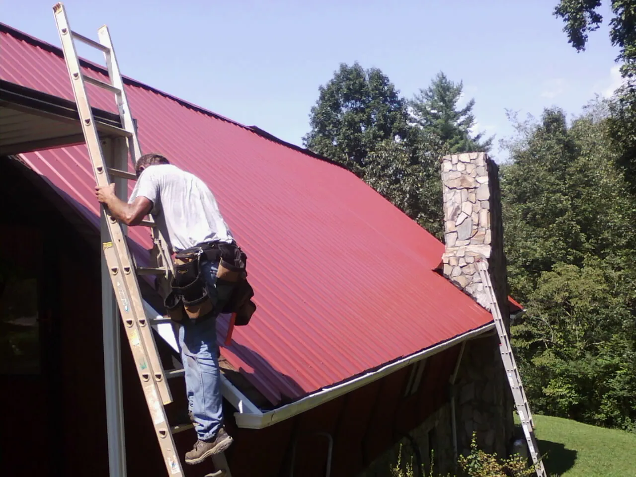 Roofer on a ladder beside a red metal roof and stone chimney — Soffit & Fascia Repair by Roofing Brothers in Cullen